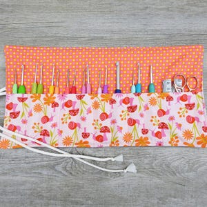 May include: A pink and orange fabric roll-up case with a white drawstring closure. The case is filled with a variety of colourful crochet hooks and a small pair of scissors. The fabric has a pattern of pink flowers and orange snails.