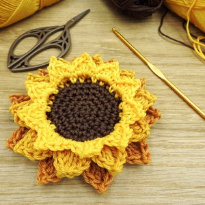 May include: A crocheted sunflower with yellow petals and a brown center. The sunflower is on a wooden surface with a crochet hook and scissors.