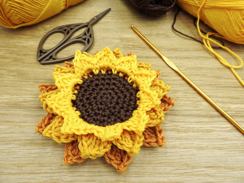 May include: A crocheted sunflower with yellow petals and a brown center. The sunflower is on a wooden surface with a crochet hook and scissors.