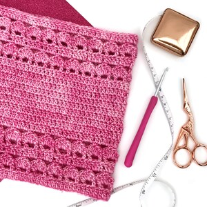 May include: A close-up of a pink crocheted fabric with a decorative edge, alongside a pink crochet hook, gold scissors, a gold square, and a measuring tape. The items are arranged on a white surface, with a glittery pink background.