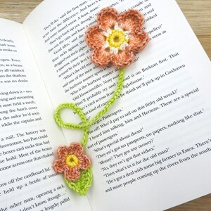 May include: A crocheted bookmark featuring two flowers. The flowers are pink and yellow with green leaves. The bookmark is placed on an open book with text about a ship and passengers.