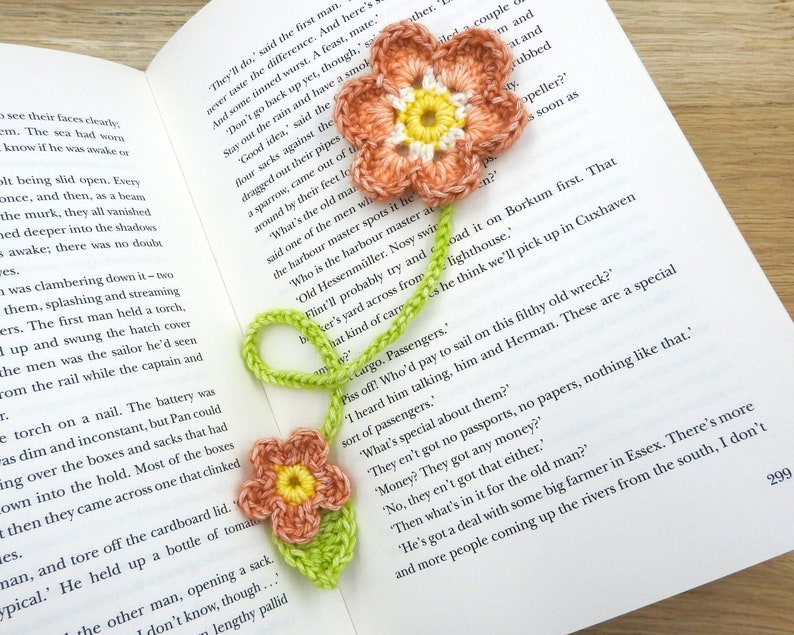 May include: A crocheted bookmark featuring two flowers. The flowers are pink and yellow with green leaves. The bookmark is placed on an open book with text about a ship and passengers.