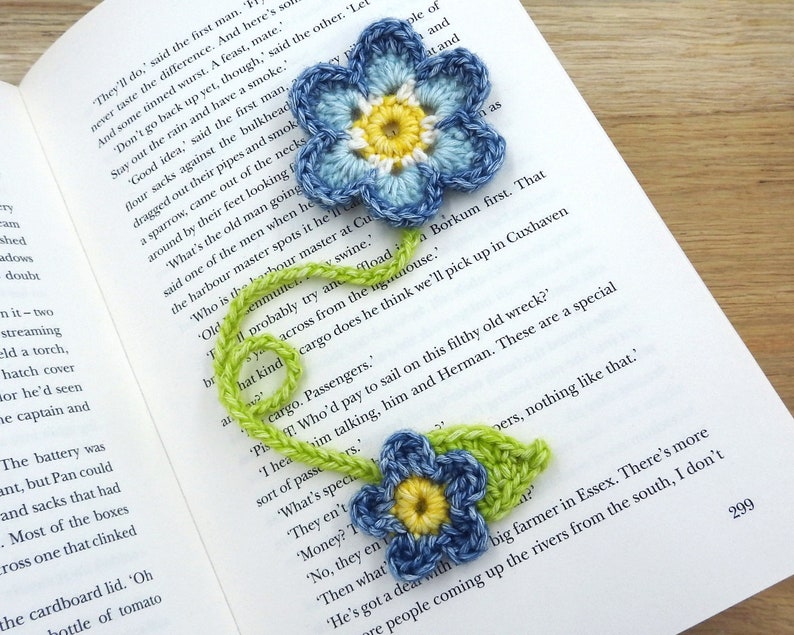 May include: Two crocheted blue flowers with yellow centres and green stems. The flowers are on a book with text.