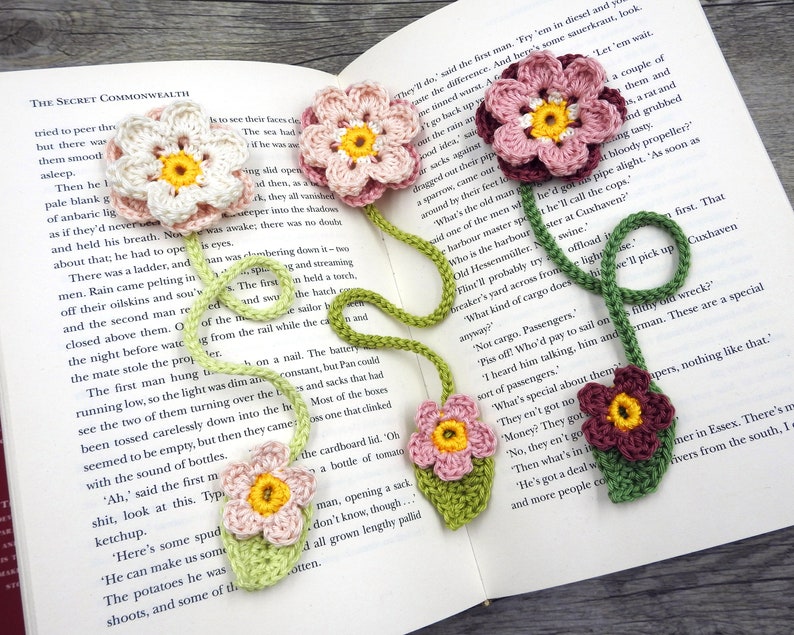 May include: A crocheted bookmark with multiple pink, red, and green flowers. The bookmark is attached to a book with the text "THE SECRET COMMONWEALTH" visible.