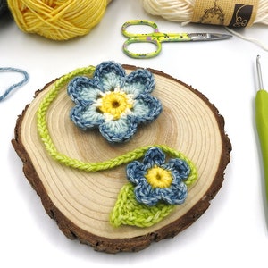 May include: Two crocheted blue and yellow flowers with green stems on a wooden slice. A green crochet hook is on the right side of the image.