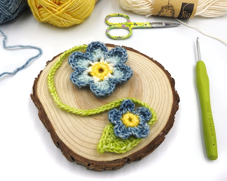 May include: Two crocheted blue and yellow flowers with green stems on a wooden slice. A green crochet hook is on the right side of the image.