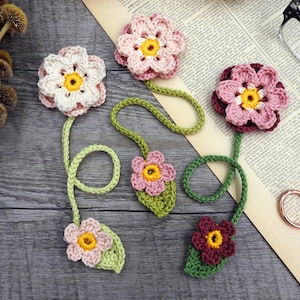 Può includere: Cinque segnalibri a uncinetto a forma di fiore con steli e foglie verdi. I fiori sono rosa, bianchi e bordeaux con centri gialli.