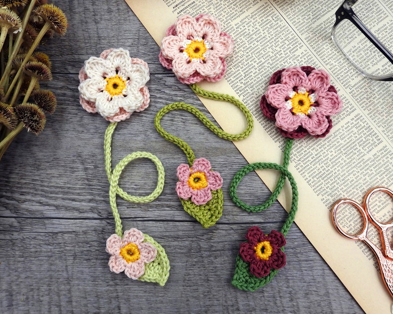 May include: Five crocheted flower bookmarks with green stems and leaves. The flowers are pink, white, and burgundy with yellow centers.