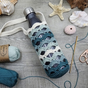 May include: A crocheted teal and white bottle holder with a grey water bottle inside. The holder is made with a crocheted strap and is sitting on a wooden surface with yarn, a seashell, and a starfish.