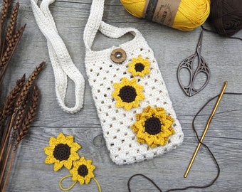 Op de afbeelding: Een witte gehaakte tas met een zonnebloemdessin. De tas heeft een lange band en is versierd met drie gele en bruine gehaakte zonnebloemen. De tas ligt op een houten oppervlak met garen en een schaar.