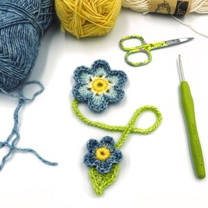May include: A close-up of two crocheted blue flowers with yellow centres, connected by a green crocheted stem. The flowers are on a white surface with balls of yarn, a crochet hook, and scissors.