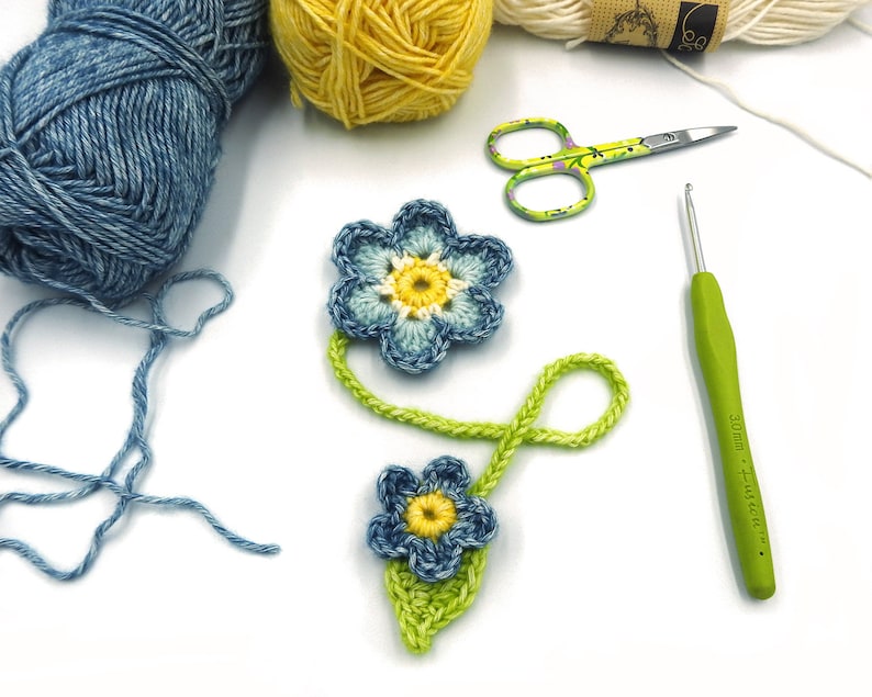 May include: A close-up of two crocheted blue flowers with yellow centres, connected by a green crocheted stem. The flowers are on a white surface with balls of yarn, a crochet hook, and scissors.