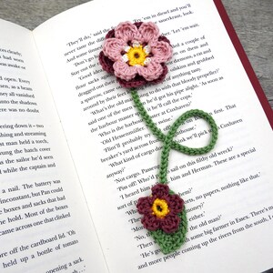 May include: A crocheted bookmark with two pink and white flowers and one burgundy flower. The bookmark is green and has a long stem. The bookmark is on top of an open book.