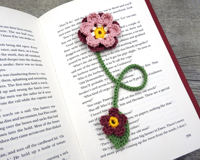 May include: A crocheted bookmark with two pink and white flowers and one burgundy flower. The bookmark is green and has a long stem. The bookmark is on top of an open book.
