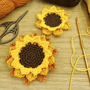 May include: Two crocheted sunflowers with yellow petals and brown centers. The sunflowers are on a wooden surface with yarn and crochet hooks.