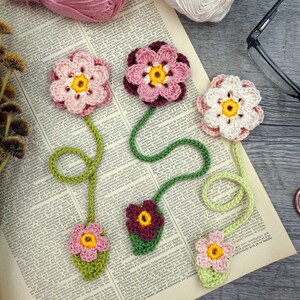 May include: Five crocheted flower bookmarks with green stems and leaves. The flowers are pink, white, and burgundy.