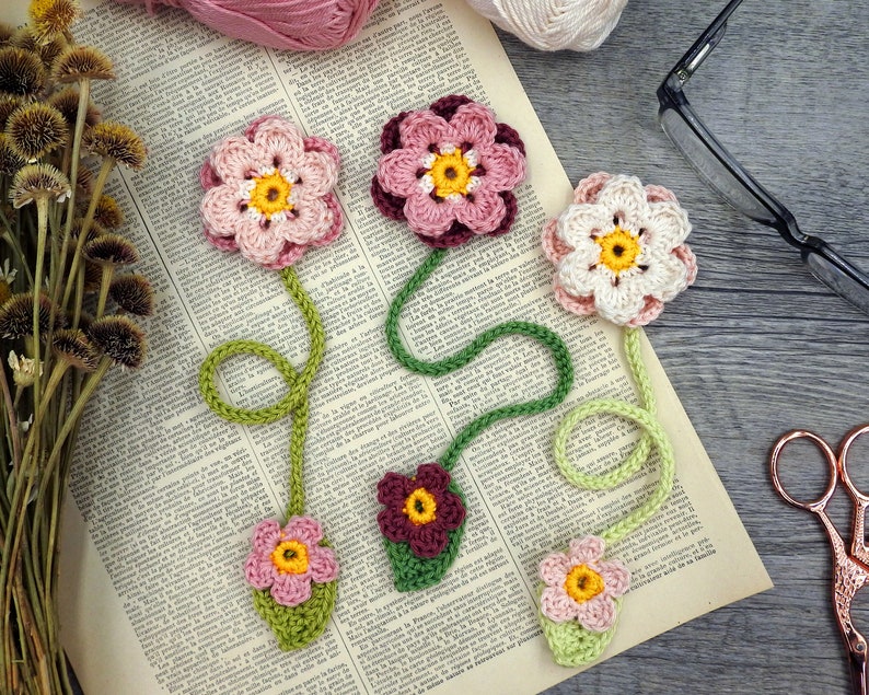 May include: Five crocheted flower bookmarks with green stems and leaves. The flowers are pink, white, and burgundy.