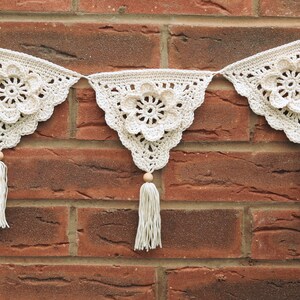 May include: A white crocheted garland with three triangular flags, each featuring a large crocheted flower. The garland is strung with wooden beads and tassels.