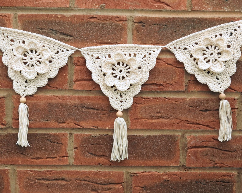 May include: A white crocheted garland with three triangular flags, each featuring a large crocheted flower. The garland is strung with wooden beads and tassels.