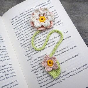 May include: A green and pink crocheted bookmark with two flower accents. The bookmark is laying on a book with text that reads "They'll do, said the first man. 'Fry 'em in diesel and you'll never taste the difference. And here And some tinned wurst. A feast"