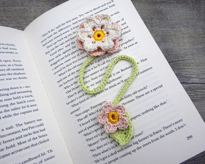 May include: A green and pink crocheted bookmark with two flower accents. The bookmark is laying on a book with text that reads "They'll do, said the first man. 'Fry 'em in diesel and you'll never taste the difference. And here And some tinned wurst. A feast"