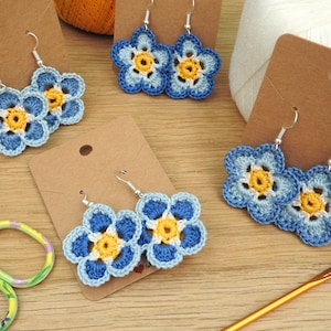 May include: Crocheted blue and yellow flower earrings with silver hooks. The earrings are displayed on a brown card.