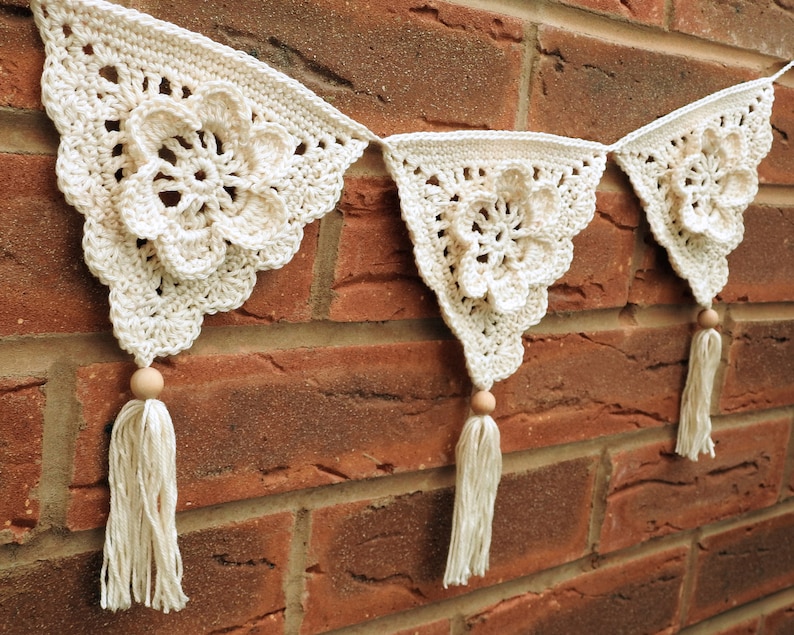 May include: A white crocheted banner with a floral design and wooden beads. The banner is hanging on a brick wall.