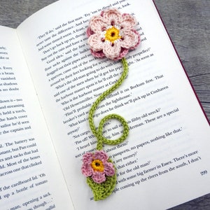 May include: A green crocheted bookmark with two pink crocheted flowers. The bookmark is shaped like a musical note and has a yellow center in each flower.
