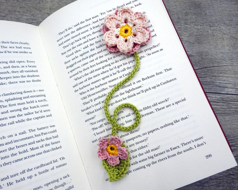 May include: A green crocheted bookmark with two pink crocheted flowers. The bookmark is shaped like a musical note and has a yellow center in each flower.