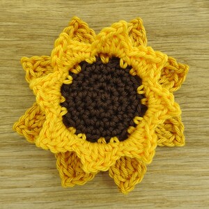 May include: A crocheted sunflower with yellow petals and a brown center. The sunflower is on a wooden surface.