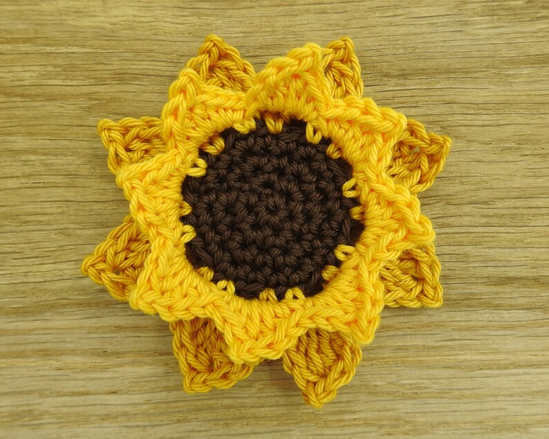 May include: A crocheted sunflower with yellow petals and a brown center. The sunflower is on a wooden surface.