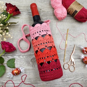 Op de afbeelding: Een gehaakte wijnfleshoes in roze en rode tinten met een hartjespatroon. De hoes ligt op een houten oppervlak met een fles wijn erin.