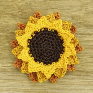 May include: A crocheted sunflower coaster with brown, yellow, and orange petals. The coaster is made of yarn and has a textured surface.