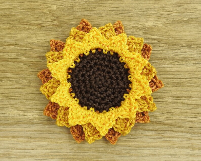 May include: A crocheted sunflower coaster with brown, yellow, and orange petals. The coaster is made of yarn and has a textured surface.