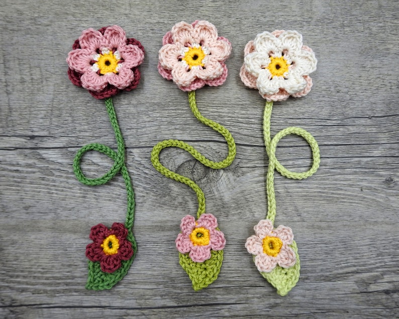 May include: Three crocheted flower bookmarks with green stems and leaves. The flowers are pink, white, and red with yellow centers.