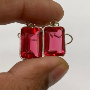 May include: A pair of rectangular earrings with a vibrant red gemstone set in a silver metal frame. The gemstones have a faceted cut, reflecting light. The earrings are held up by a silver hook.