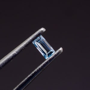 Può includere: Una pietra preziosa rettangolare con taglio baguette, di colore azzurro e superficie sfaccettata, tenuta da una pinzetta metallica. La pietra mostra riflessi interni e trasparenza, mettendo in risalto una tonalità blu chiara e fredda.