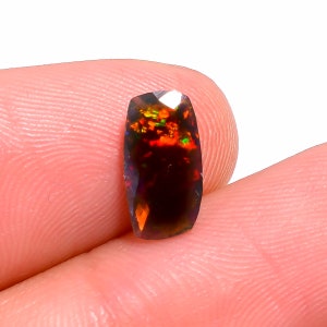 May include: A faceted, rectangular black opal gemstone with vibrant internal fire. The opal displays flashes of red, orange, and green. The gemstone is held between two fingers, showcasing its polished surface and unique color play.