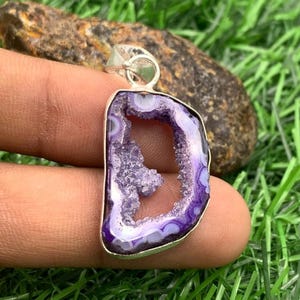 Puede incluir: Un colgante que presenta una rebanada de ágata morada con un centro de cristal druzy, con forma de letra D. La piedra está engastada en un marco de metal plateado con una anilla en la parte superior, mostrando bandas naturales y texturas cristalinas.