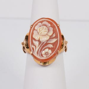 May include: Oval cameo ring featuring a carved floral design in shades of orange and cream. The ring has a gold-colored band with intricate detailing around the cameo and on the sides. The ring is displayed on a white stand.