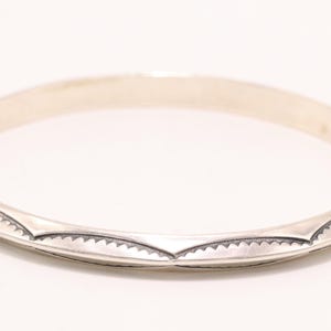 May include: A silver-colored bangle bracelet with a decorative pattern. The bracelet has a smooth, rounded surface with a series of etched, triangular designs along the edges. The bracelet is circular and appears to be made of metal.