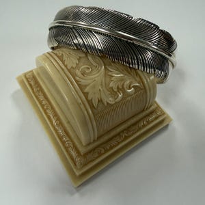 May include: A silver cuff bracelet shaped like a large feather with detailed engraved lines along the surface. The bracelet is resting on a cream-colored, rectangular display stand featuring embossed floral patterns and ribbed textures.
