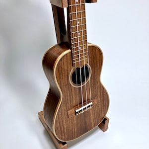 May include: A brown ukulele with a dark wood grain finish, resting on a matching wooden stand. The instrument has a round sound hole and four strings. The stand is designed to hold the ukulele upright.