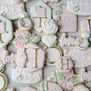 Pu&ograve; includere: Una collezione di biscotti decorati a tema matrimonio, con glassa rosa e bianca, scritte dorate, accenti floreali, forme di anelli con diamante e testi come Bride to be, love, e varie date come 03 03 22 e 06 03 22.