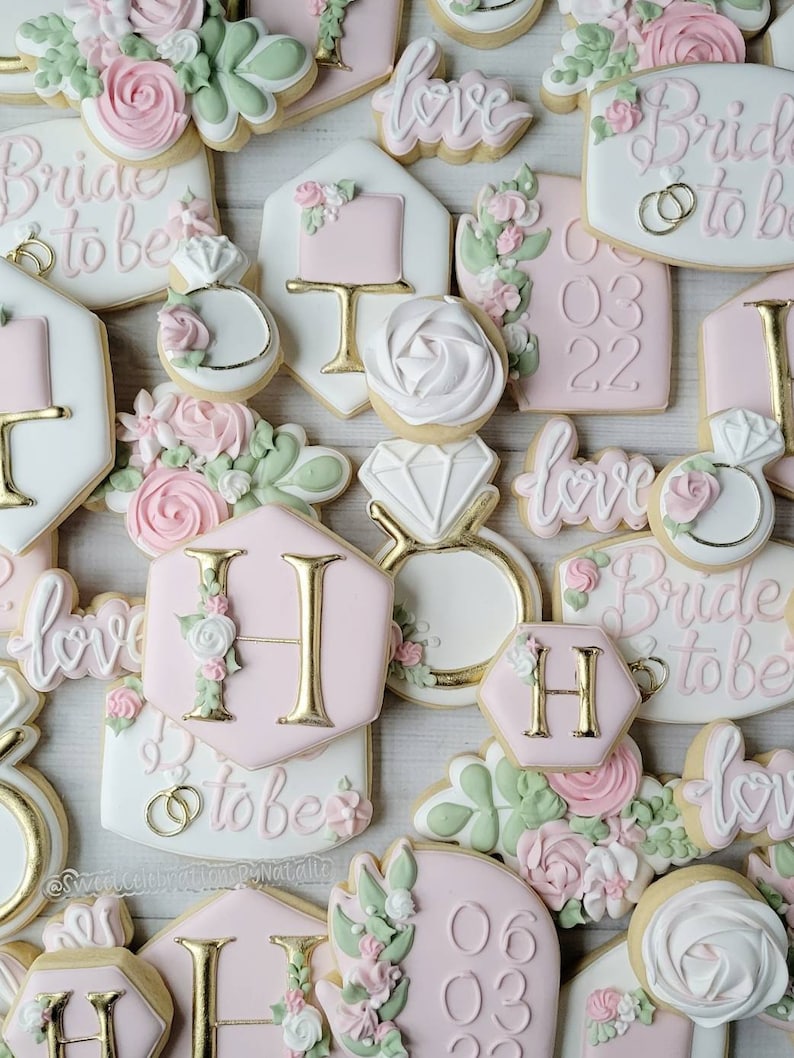 Pu&ograve; includere: Una collezione di biscotti decorati a tema matrimonio, con glassa rosa e bianca, scritte dorate, accenti floreali, forme di anelli con diamante e testi come Bride to be, love, e varie date come 03 03 22 e 06 03 22.