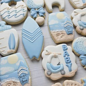 Puede incluir: Una colecci&oacute;n de galletas decoradas con motivos de beb&eacute;s y surf en tonos azul claro, blanco y beige. Los dise&ntilde;os incluyen tablas de surf, bodies con el texto crawl. walk. surf., biberones y escenas de playa con huellas en la arena.