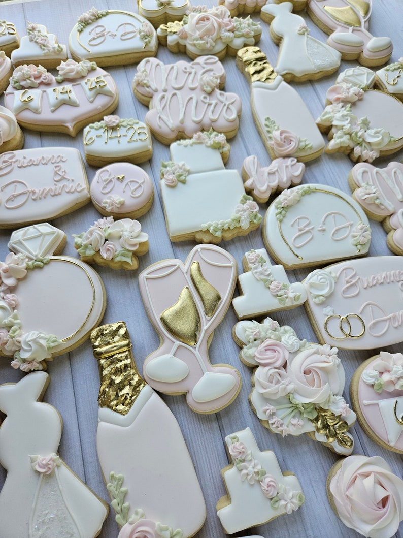 Pu&ograve; includere: Una collezione di biscotti a tema matrimonio decorati con glassa bianca e rosa pallido, accenti dorati e dettagli floreali. Le forme includono bottiglie di champagne, abiti da sposa, anelli, torte e targhe con testo come Brianna & Dominic.