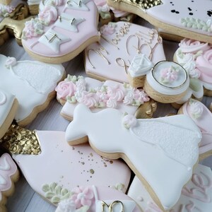 Pu&ograve; includere: Una collezione di biscotti decorati a tema matrimonio, tra cui forme di abiti bianchi, anelli di fidanzamento e bottiglie di champagne. I biscotti sono glassati in tonalit&agrave; rosa pallido, bianco e oro, con la scritta Marissa & Billy.