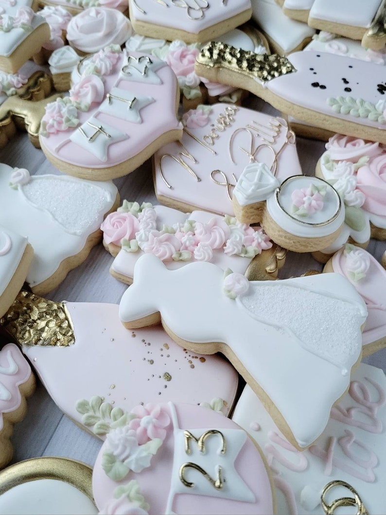 Pu&ograve; includere: Una collezione di biscotti decorati a tema matrimonio, tra cui forme di abiti bianchi, anelli di fidanzamento e bottiglie di champagne. I biscotti sono glassati in tonalit&agrave; rosa pallido, bianco e oro, con la scritta Marissa & Billy.