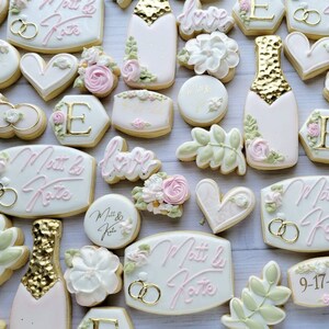 Pu&ograve; includere: Una collezione di biscotti decorati con bottiglie di champagne con foglia d'oro, fiori bianchi, foglie verdi, cuori e targhe esagonali. Diversi biscotti mostrano il testo Matt & Kate e la data 9-17-22 in glassa rosa e dorata.
